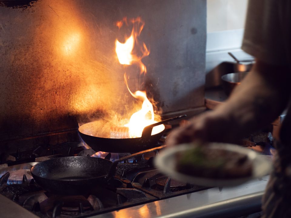 A pan flaring up over the gas range in Tusco's kitchen, with a plated dish being carried past